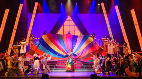 The ensemble performing Joseph at the Bournemouth Pavilion. There are groups of people on the left and right of the stage with a woman in a rainbow coat with a enormous rainbow cape as well.