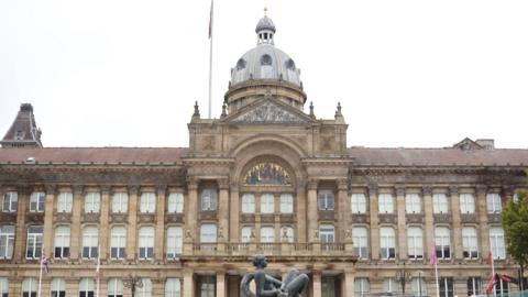 The exterior of the Birmingham City Council building, a Victorian-style stone building with a large central dome atop the pillared and arched entrance. There are wide steps and a fountain with a black sculpture of a person in front of it.