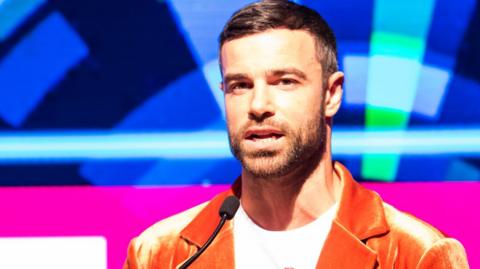 Ben Starr speaking on stage, wearing an orange jacket