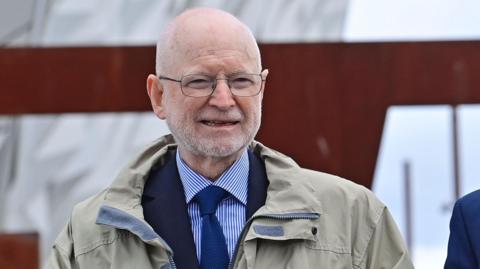 Sir Desmond Rea, a man with a shaved head and a short, white beard, walks towards the camera outside the Titanic Belfast building in 2022. He is wearing glasses, a grey/beige anorack over a navy suit jacket, blue and white shirt and a navy tie. 