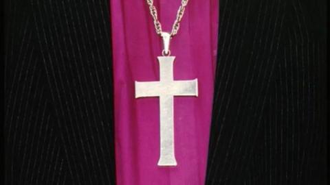 A cross on a purple garment, there is black clothing on either side.