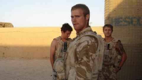 Three people including Mark Omrod in desert camouflage gear standing on sandy ground near a wall and cylindrical structure outdoors.