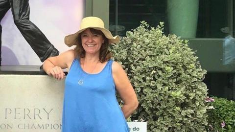 Henrietta Harrison smiles at the camera as she is photographed in a blue dress and straw hat stands. She is stood in front of some bushes and a statue. 