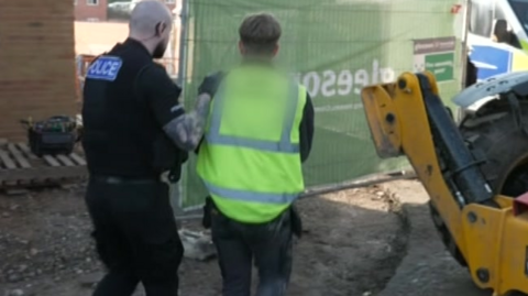 The suspect, who has brown hair, is wearing a florescent vest over clothing as he is walked off a building site by an officer in uniform. 