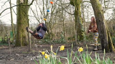 A little girl is swinging in a forest on a swing in front of a patch of daffodils