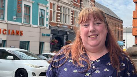 Emma Walton standing on a street in a town centre beside a brick building, with shops, parked cars and historical buildings visible behind her.