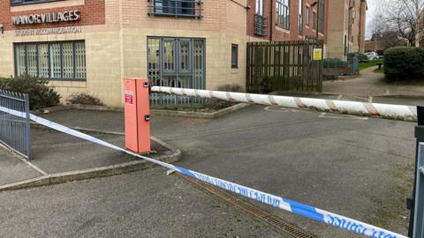 Blue and white police tape is in place across the entrance to a car park. A red and white car park barrier is behind the police tape.