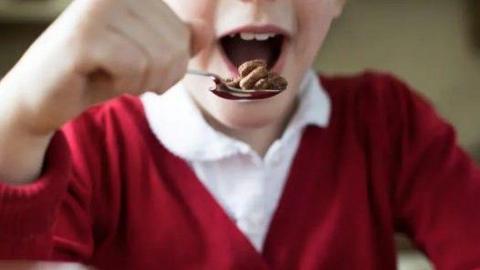 A child wearing a burgundy jumper and white shirt lifts a spoonful of cereal up to his mouth