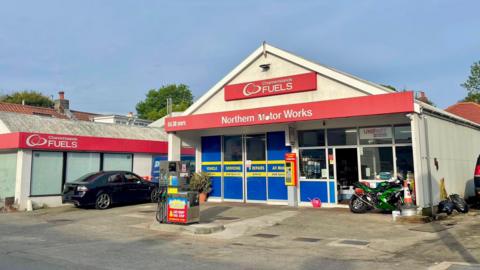 The image shows a small fuel station and motor works shop with a white and red signage. The main sign reads "Northern Motor Works" There is a black car and a green motorcycle  parked near the entrance, along with some other items like tires and equipment. 