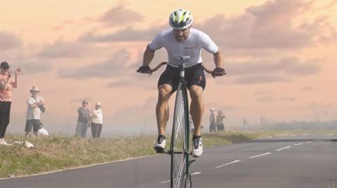 A man in cycling gear on a penny farthing. He is on a road with people on the side of the road clapping him on.