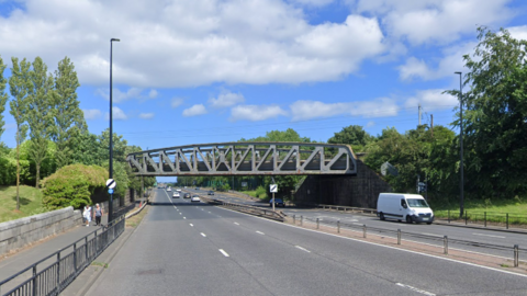 A three-lane dual carriageway with a footbridge above it. There are national speed limit signs at the side.