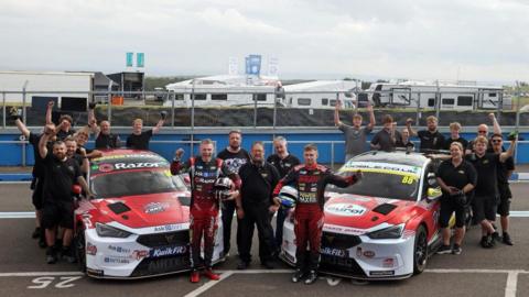 A group of people stand on a racing track next to two black, white and red racing cars which are covered in various logos. 