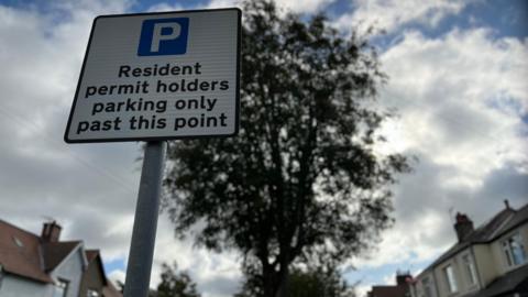 A close up of a blue and white 'residents permits holders only' parking sign, with homes and trees in a pleasant street in the background