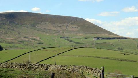 View of the green Pendle Hill on a sunny day under a blue sky. Low green fields can be seen in the foreground with a stone wall at the bottom image.
