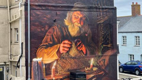 A mural on the side of a building depicts an older man with a long white beard, wearing a hat and smoking a pipe, as he mends a fishing net. He is illuminated by two candles in what appears to be a workshop or cabin