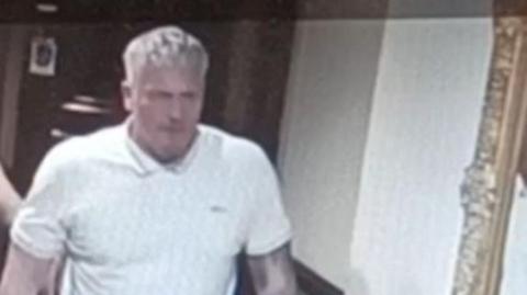 A standing man with blonde hair. He is wearing a white polo shirt and has a tattoo sleeve on his left arm.