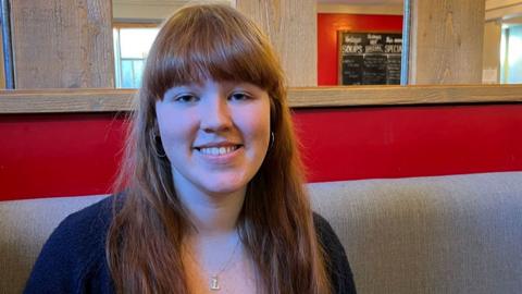 Latonya Skye-Patterson, a 20 year old woman with long, auburn hair sits in a cafe looking at the camera