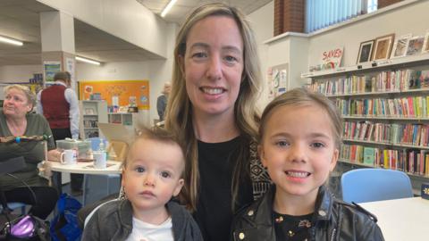 A woman with blonde hair smiling at the camera. She is wearing a black t-shirt and black-and-white patterned jacket. On the left of the picture, a baby boy is looking at the camera. He is wearing a white top and a black jacket. On the right, a young girl is smiling at the camera. She has brown hair and is wearing a black jacket and a black floral top. They are in a library with books in the background. There are other people sitting and standing behind them.