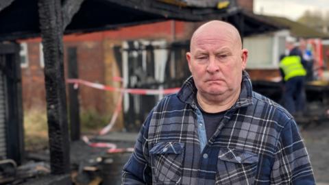 Stan Yannetta standing in front of the burnt remains of his Halloween display and a badly burnt garage. He is a middle-aged man with no hair. He is wearing a blue check shirt and looks unhappy.