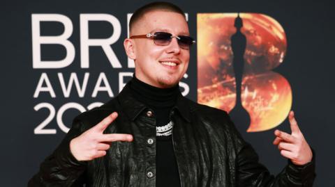 Aitch, with short brown and dark shades wearing a dark polo neck swearer and black leather jacket poses on the red carpet during the BRIT Awards at the Co-op Live Arena, in Manchester, Britain, 28 February 2026.