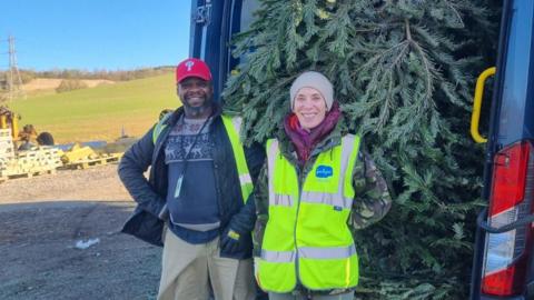 Two volunteers, a man and a woman, stand in front of a van, which has its boot door open and is stuffed with a Christmas tree. There are wooden pallets in the background and rolling, green fields. It is a sunny day.