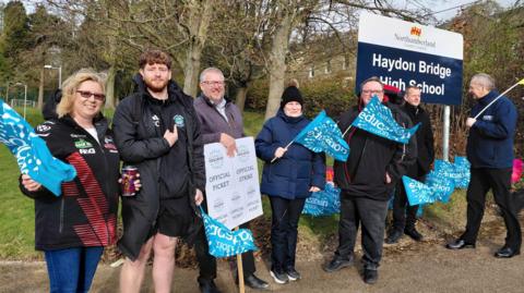 Education workers and supporters holding National Education Union flags and an official strike picket sign outside Haydon Bridge High School. There are seven strikers standing next to a sign at the entrance to Hayden Bridge High School.