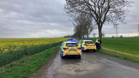 The road is a narrow one with no road markings with fields either side, one with flowering oil seed rape. Two police cars are parked across the road to stop traffic driving down it. Gravel or sand lays on the surface in front of them. Two police men stand next to a tree.