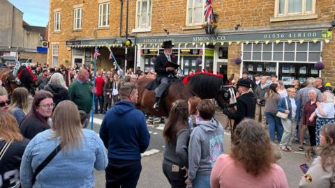 A man in a top hat sit on a horse and it reading a proclamation in front of a crowd of people in  town street
