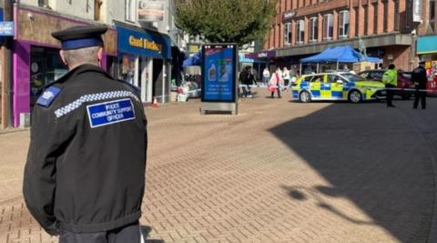 Shops and buildings can be seen either side of the main pedestrian route on Parliament Row. A police car and cordon is visible and shoppers stop to look at the scene. A police officer is in the foreground watching the scene.
