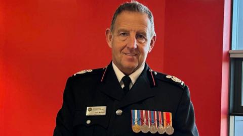 Jim McNeil smiles as he looks directly into the camera. He has short grey hair and is wearing his fire officer's uniform - a black blazer adorned with five medals on the left. The wall behind him is red.