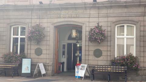 The front of the Jersey Town Hall. The wooden door is open inwards. There are glass automatic doors after the entryway. There are foldable signs around the doorway. A bench either side of the door. Above the door is written Salle Paroissiale De St Helier. The are pink flowers hanging either side of the doorway and in flowerbeds in front of the two windows, one either side of the door. 