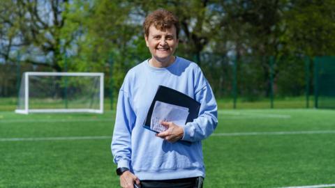 Julie Norris with short brown hair, blue jumper and black clipboard smiling and while standing on a football pitch. There is a goalpost and some trees in the background.