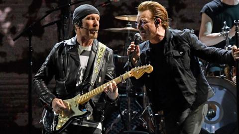 The Edge in black leather jacket and grey hat, playing a guitar, alongside Bono singing into microphone.