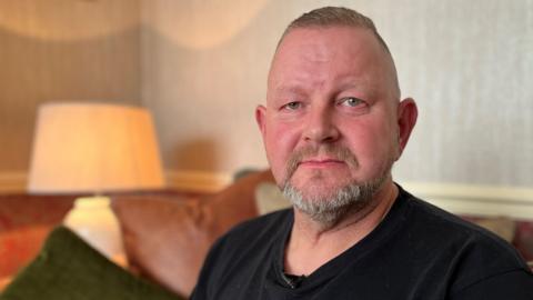 A man wears a serious expression as he sits in a cosy-looking lounge. He has cropped light-brown hair and a short salt-and-pepper beard. He wears a black T-shirt.