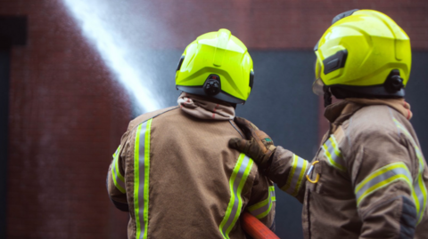 Two firefighters, in uniform, have the back to the camera. The firefighter on the left is holding a hose, which is spraying water. The one on the right has their hand resting on the shoulder of the other firefighter
