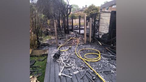 A garden with a severely burned out structure in the centre. Wooden decking in the foreground is severely charred.