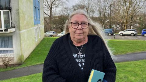 A woman wearing glasses, a cross necklace and holding a book stood in front of a building. There is an area of grass and a number of cars parked on the road behind her. 