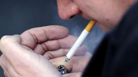 A close up of a smoker holding a lighter in one hand with ash hanging from a cigarette which is alight and in their mouth.