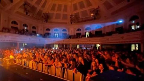 A crowd of people look at a stage in a large hall. There are spotlights shining above their heads.