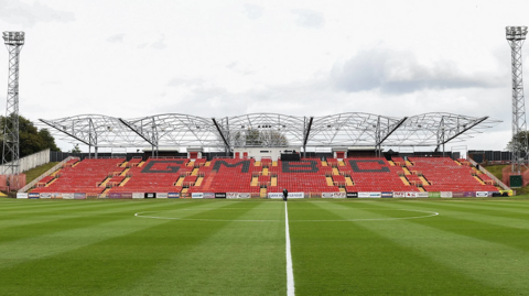 A picture showing the grass pitch at Gateshead International Stadium. The picture is taken from the half-way line looking across the pitch. On the other side is a stand full of red seats. A number of black seats spells out GMBC.