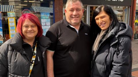 Two women and a man are standing on a high street facing the camera. The man is in the middle and wearing a black Hugo Boss T-shirt. The woman on the left has red hair and a black coat. The woman on the right has dark hair, a light scar and a black coat.