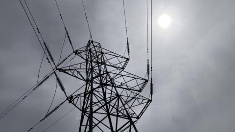 An electricity tower pictured silhouetted against a grey sky in Inverness. The structure is tall with metal lattice work and cables attached to it.