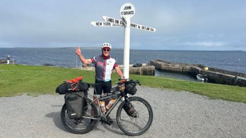 Norman Brown with his one thumb up wearing sunglasses and a helmet holding his bike next to the John O'Groats landmark. The sea is in the background.