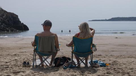 The backs of two people sat on deckchairs. One is a topless man wearing a baseball cap and sunglasses with a towel on the back of his blue chair. The other is a woman with long blonde hair in a red swimsuit, sat in a matching blue chair with one hand up to shield her eyes from the sun. The beach is sandy, and they have bags and shoes beside them and are looking out at the sea where two more people are swimming. There are cliffs on either side. 