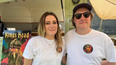 A head and shoulders image of a woman and a man wearing white T-shirts with the King's Head logo on them. They are standing in front of a mobile bar and next to a large poster of The King's Head logo, which depicts a Viking-type character under the words King's Head.