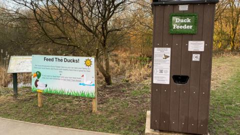 A duck vending machine stands next to an information board about feeding ducks in a country park