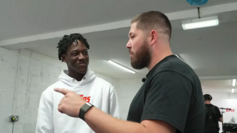 Manchester United footballer Kobbie Mainoo is on the left hand side speaking to John Nellis on the right. Mainoo is wearing a white hoodie with red lettering. Nellis is in a black t-shirt and has a beard and short, shaven hair