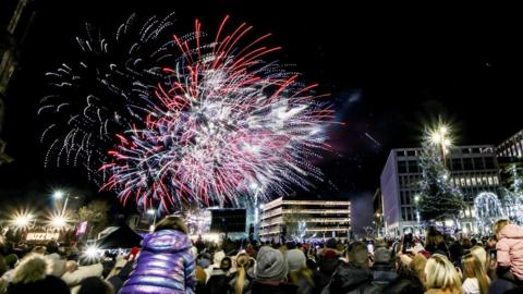 Fireworks explode in the night sky as crowds look on in Keel Square. A Christmas tree is illuminated in the right-hand side of the image alongside a number of other festive decorations. Many people in the crowd are wearing coats and woolly hats.