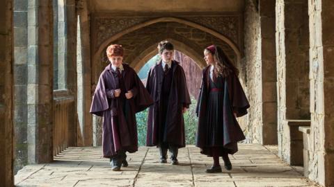 Alastair Stout as Ron Weasley, Dominic McLaughlin as Harry Potter and Arabella Stanton as Hermione Granger in the HBO YV series walking along a cloister of fictional Hogwarts school set. 