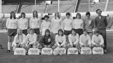 A black and white image of a group of women in football kit. They are stood in a football stadium.